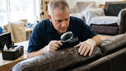 Furniture technician examining a tear in dark leather sofa upholstery using a magnifying glass.