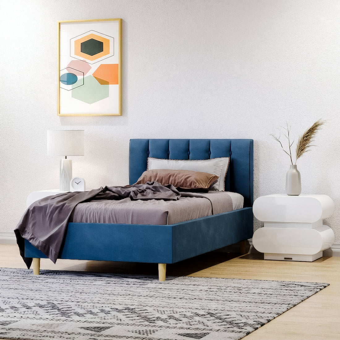 Modern guest bedroom featuring a navy blue upholstered single bed frame with a vertical channeled headboard. The room includes white sculptural "capsule" style nightstands, a geometric abstract wall art piece in orange and green, and a grey patterned rug over light wood floors.