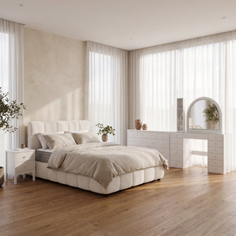 Modern neutral-toned Airbnb bedroom featuring a cream boucle upholstered bed dressed in beige linens, a matching white nightstand, and a coordinated dresser and arched vanity setup against floor-to-ceiling sheer curtains.