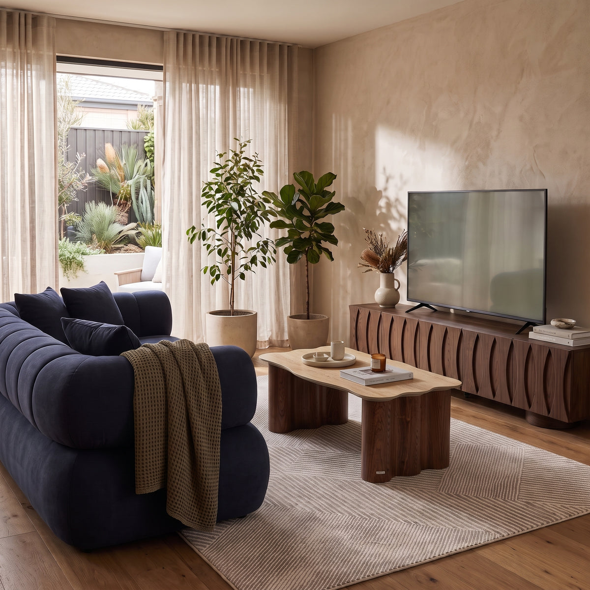 Navy blue tufted sofa paired with a wavy timber coffee table and a dark wood TV unit in a sunlit living room setting.