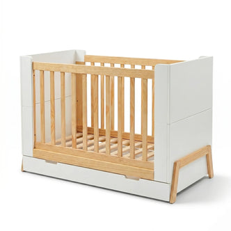 A modern baby crib featuring a white frame with natural wood slats and angled light-wood legs, including a storage drawer at the base.