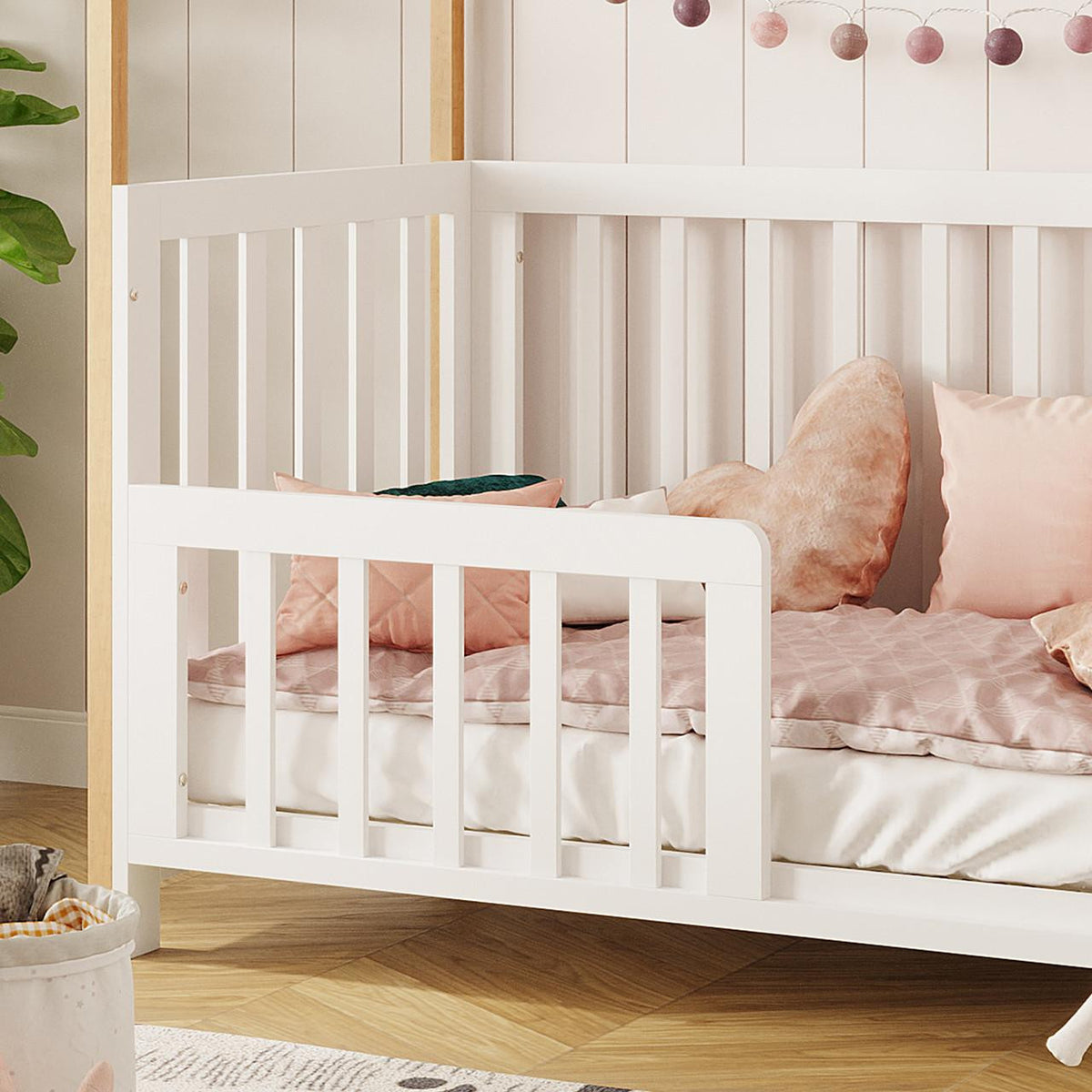 Damaris & Giro Solid Pine Baby Cot with Convertible Rail Set White, featuring a white wooden frame with vertical slats, dressed in soft bedding, surrounded by cozy pillows and a string of ambient lights.