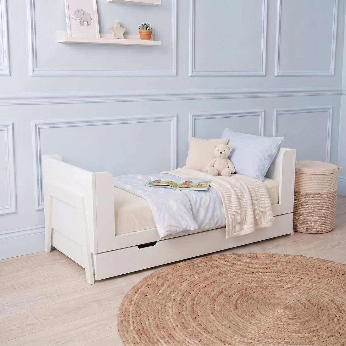 White baby cot with pull-out storage drawer, side rails, and mattress with light blue bedding in nursery setting