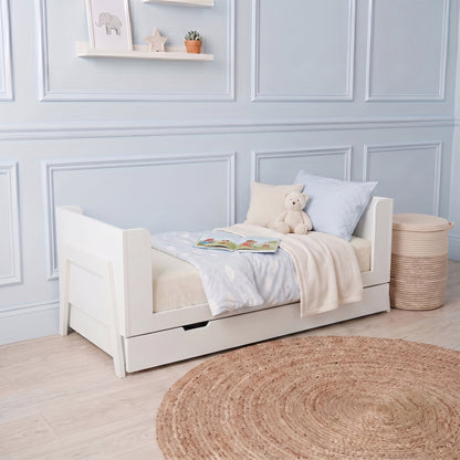 White baby cot with pull-out storage drawer, side rails, and mattress with light blue bedding in nursery setting
