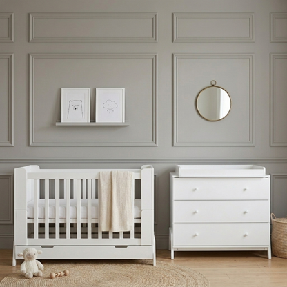 White baby cot with pull-out storage drawer and vertical slats, shown beside white chest of drawers in grey panelled nursery