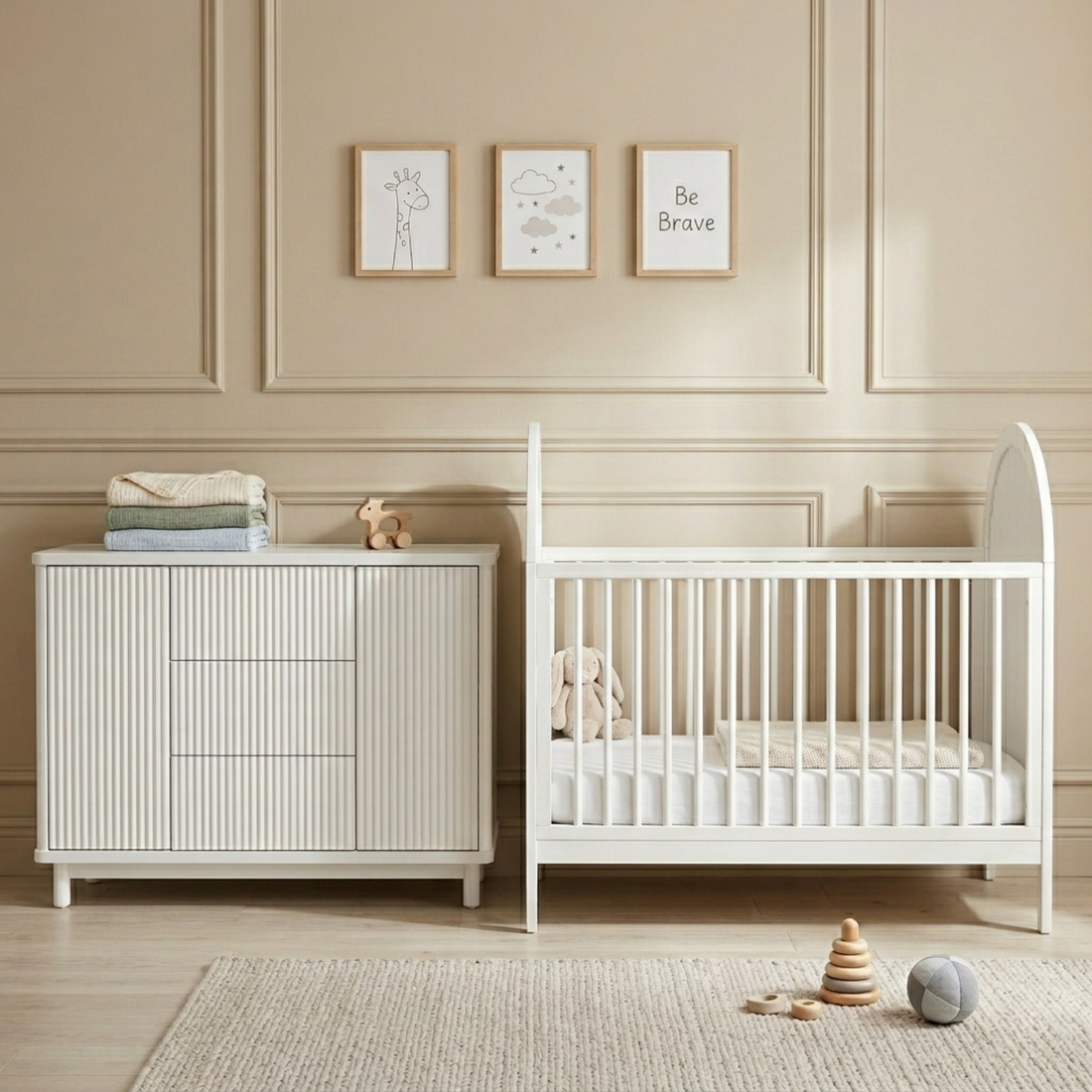 White baby cot with arched headboard and vertical slats beside ribbed chest of drawers, wooden toys on textured rug
