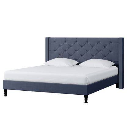 Maggie King Winged Fabric Bed Frame – Dark Blue features a deep blue, button-tufted headboard, platform base, and black legs, accompanied by a white mattress and pillows, embodying sophistication and grandeur.