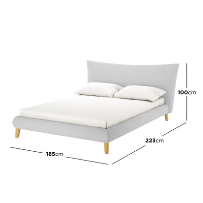Silvina Queen Upholstered Boucle Bed in Light Grey features a low-profile design, flared headboard, and tapered wooden legs. The bed is seen with a white mattress and two pillows, showcasing its elegant structure.