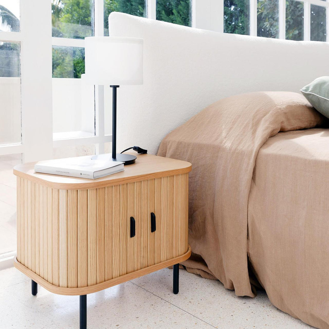 Aaron 50cm Bedside Table with Sliding Doors - Natural, featuring vertical slats, slim black legs, topped with a minimalist lamp and book, set in a light-filled bedroom corner near a bed.