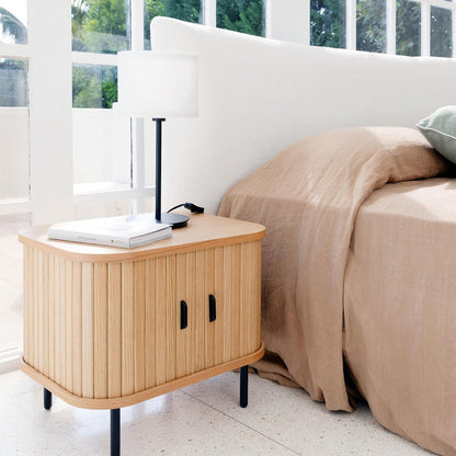 Aaron 50cm Bedside Table with Sliding Doors - Natural, featuring vertical slats, slim black legs, topped with a minimalist lamp and book, set in a light-filled bedroom corner near a bed.