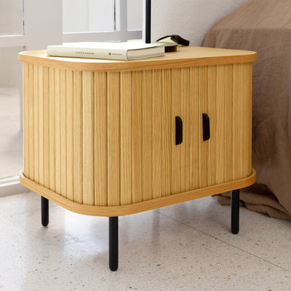 Aaron 50cm Bedside Table with Sliding Doors - Natural features light-stained wood, vertical slatted doors, and slender black metal legs. Topped with a book titled “Copenhagen” and a small lamp base.
