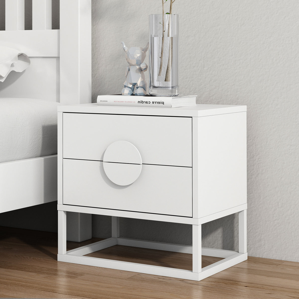 Abigail 55cm 2-Drawer Bedside Table w/ Handle - White beside a bed, featuring arc handles, atop a metal base, with a book, geometric bear figurine, and vase on its surface.