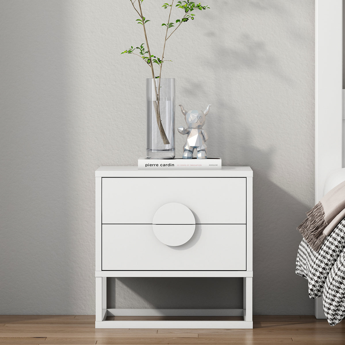 The Abigail 55cm 2-Drawer Bedside Table in white features sleek arc handles, topped with a glass vase, pierre cardin book, and silver bear figurine, complementing a minimalist bedroom setting.