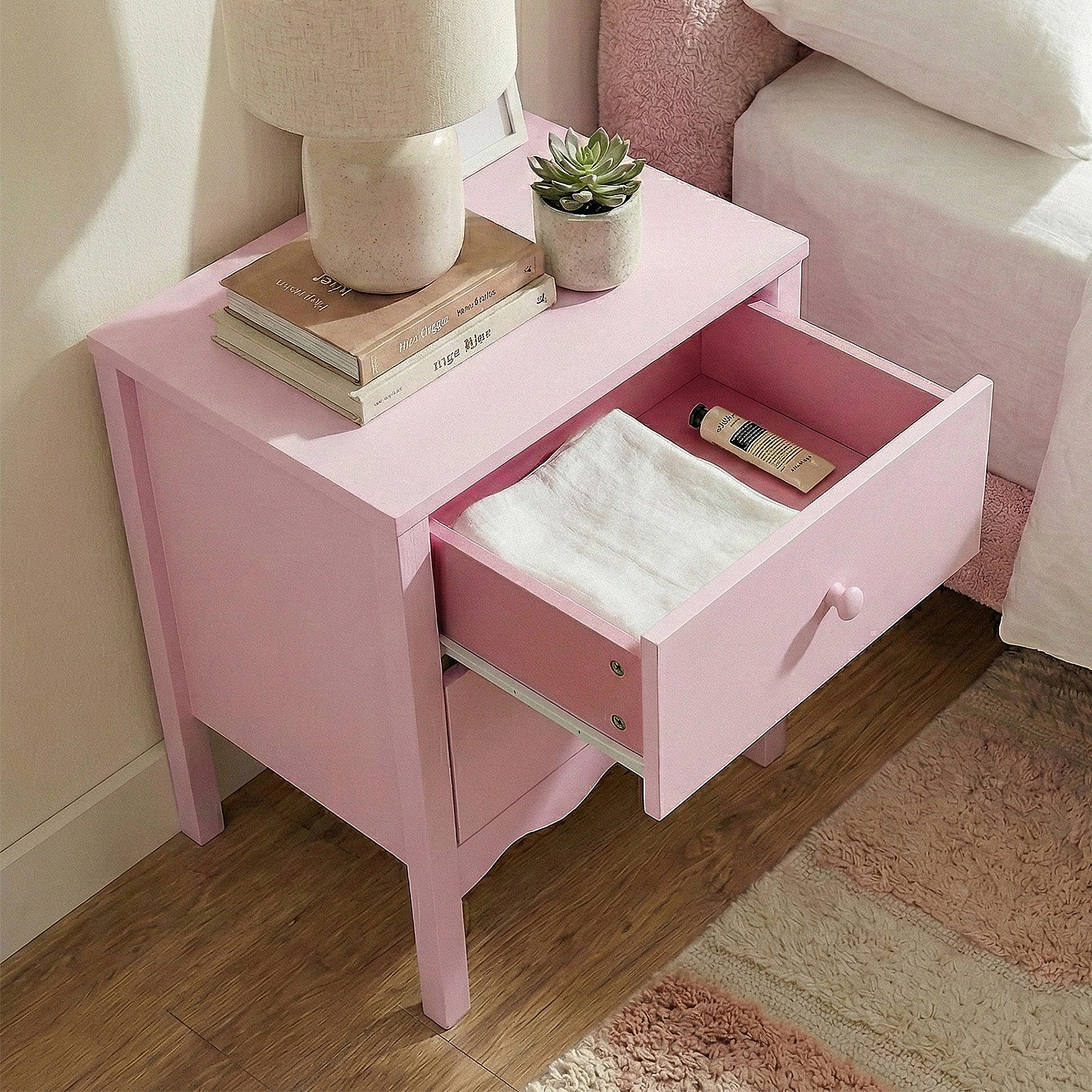 Pink two-drawer bedside table with open top drawer showing white towel and tube, styled with lamp, books, and succulent plant