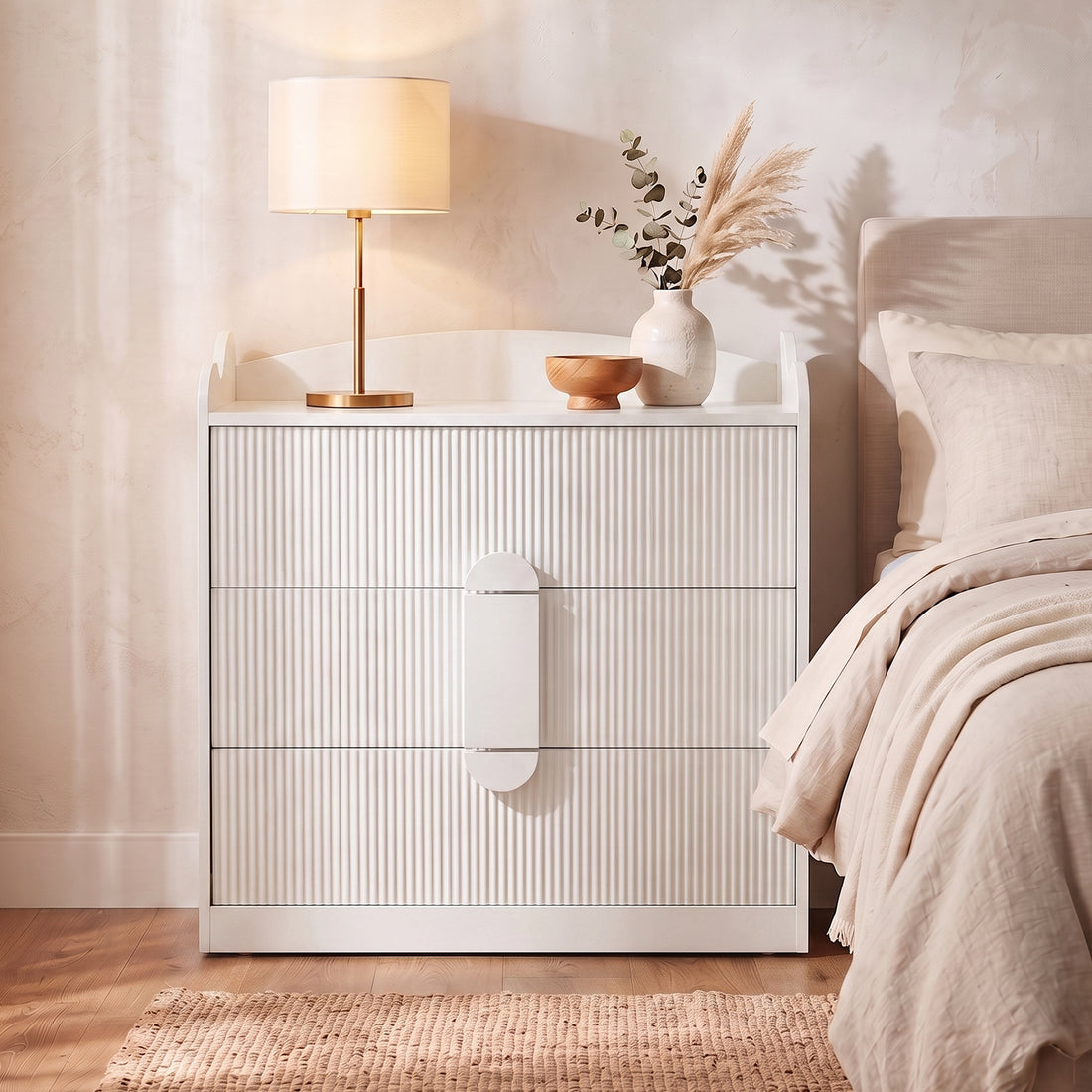 White fluted three-drawer chest with rounded handle detail in a bedroom setting beside a bed with beige linens, styled with a
