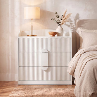 White fluted three-drawer chest with rounded handle detail in a bedroom setting beside a bed with beige linens, styled with a
