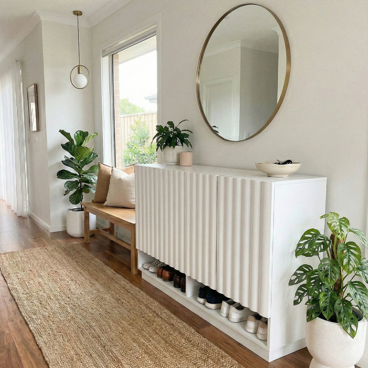 White fluted shoe cabinet with open shelf displaying shoes, round mirror above, beside wooden bench with cushions and indoor
