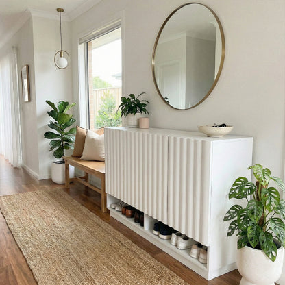 White fluted shoe cabinet with open shelf displaying shoes, round mirror above, beside wooden bench with cushions and indoor