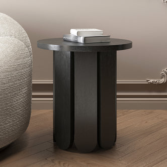 Hugo 45cm Round Side Table with Curve Detail in Black, featuring a scalloped-edge base, is placed on a wood-plank floor beside a boucle-style chair, topped with two stacked books.