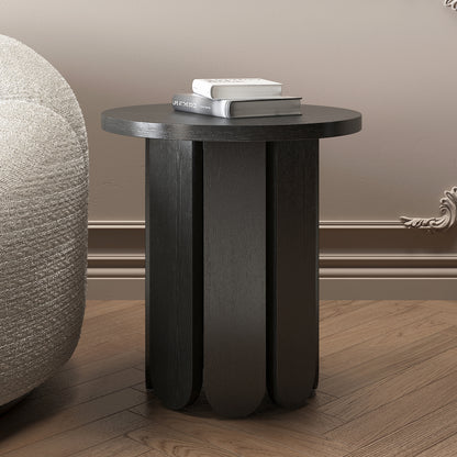 Hugo 45cm Round Side Table with Curve Detail in Black, featuring a scalloped-edge base, is placed on a wood-plank floor beside a boucle-style chair, topped with two stacked books.