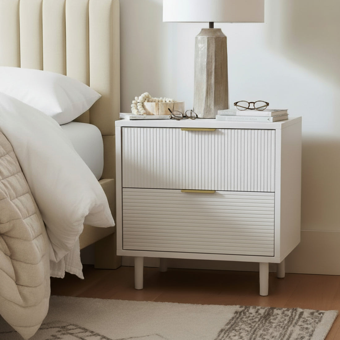Zuriel 45cm Bedside Table with Drawers in White next to a bed, featuring a faceted lamp, books, wooden bowl, and eyeglasses, emphasizing its sleek design and practical storage.