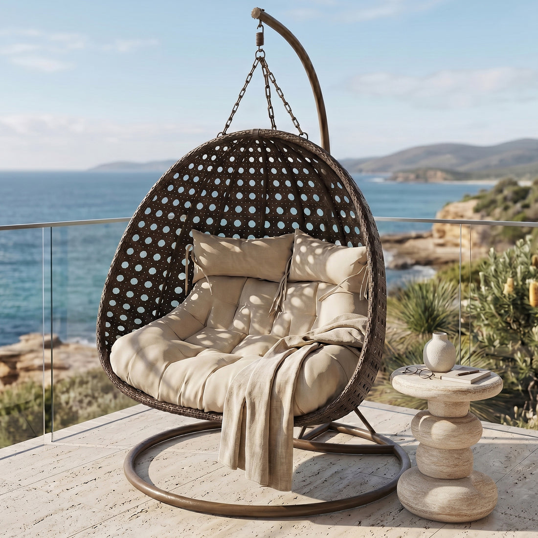 Buena 2-Seater Hanging Egg Chair in brown wicker with beige cushions and throw blanket, suspended from arched metal stand