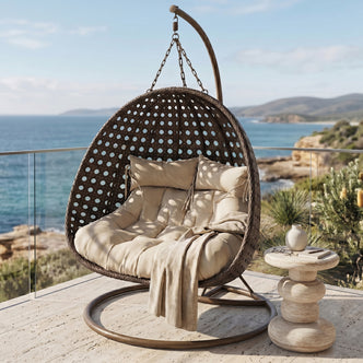 Buena 2-Seater Hanging Egg Chair in brown wicker with beige cushions and throw blanket, suspended from arched metal stand