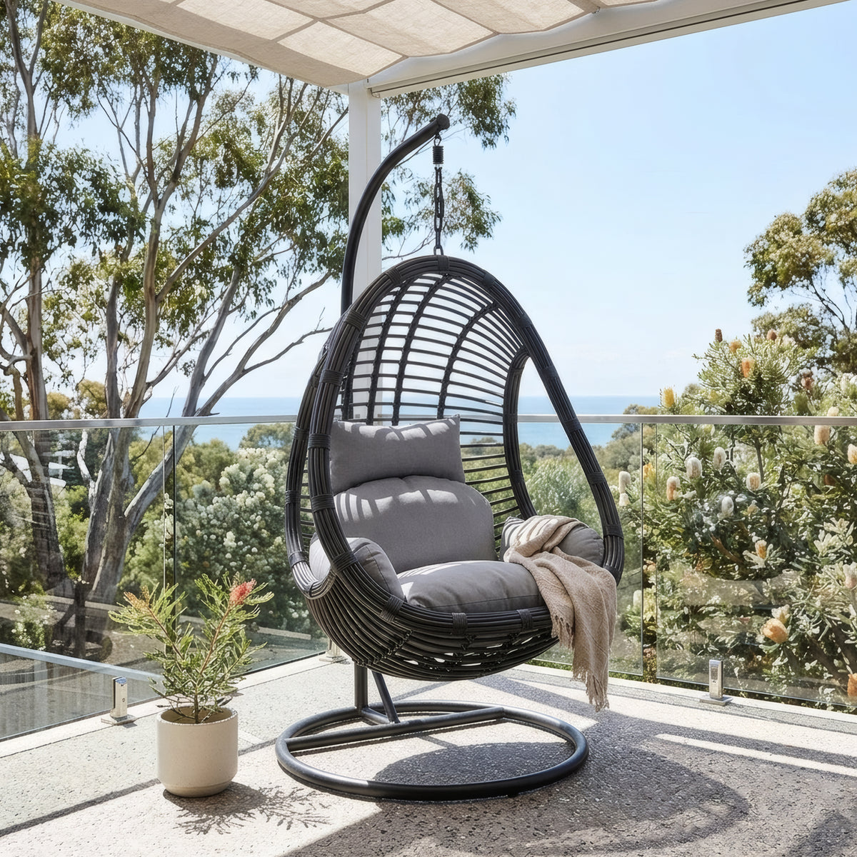Deanna Hanging Egg Chair in black rattan with grey cushions and curved metal stand on outdoor balcony with ocean view