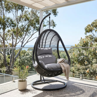 Deanna Hanging Egg Chair in black rattan with grey cushions and curved metal stand on outdoor balcony with ocean view