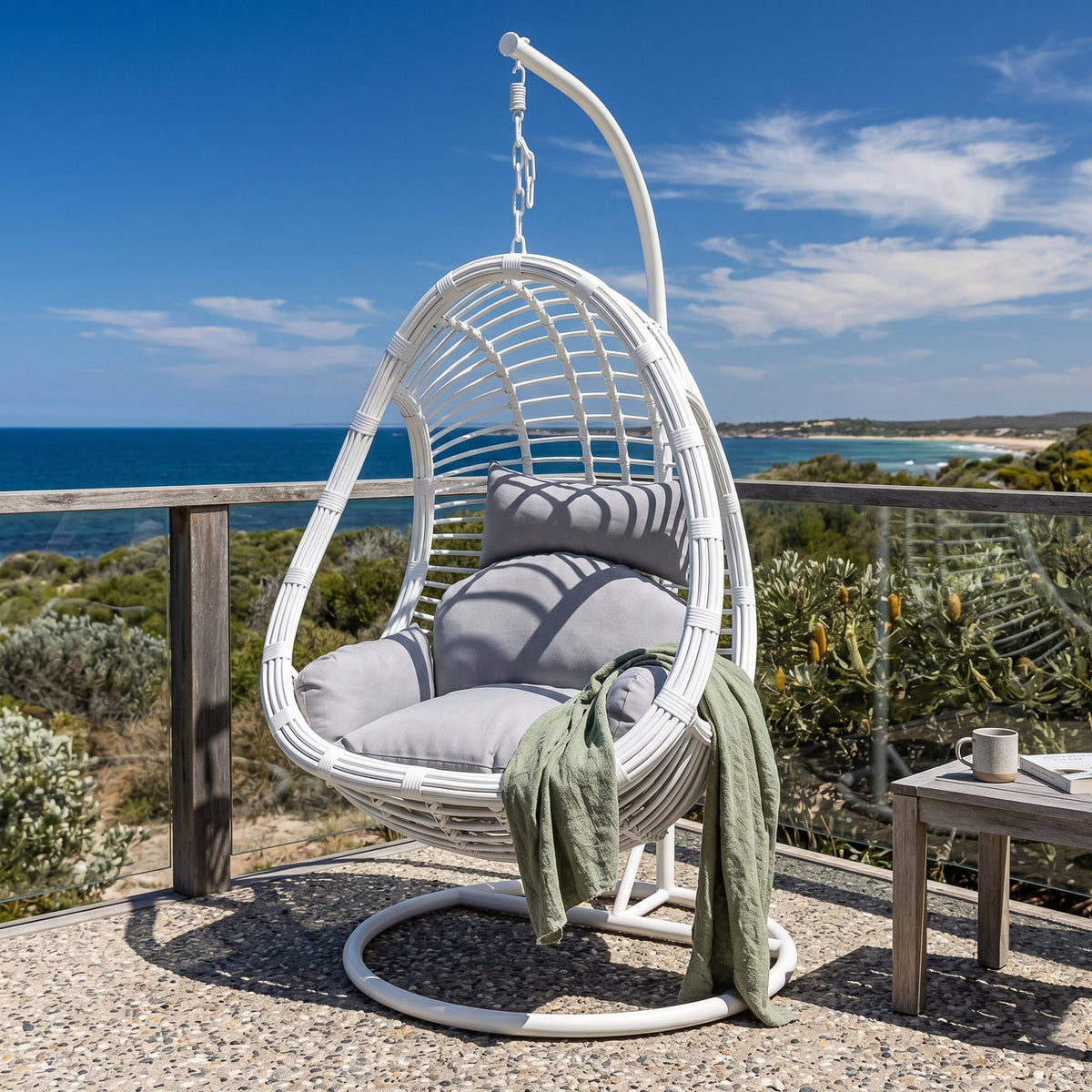 Deanna Hanging Egg Chair in White with grey cushions and green throw, on balcony with ocean view