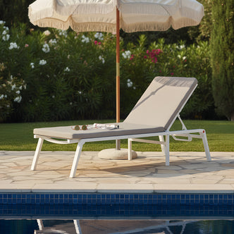 White aluminum outdoor sun lounger with a grey performance fabric cushion, positioned poolside under a cream fringed umbrella.