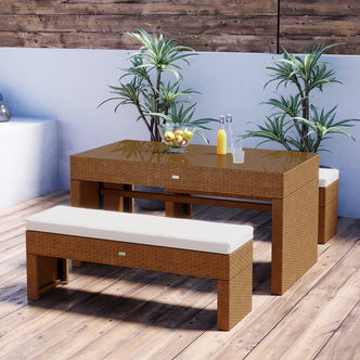 Kummi 4 Seater Outdoor Dining Bench Set features a wicker table with a glass top, two matching benches with cushions, a fruit bowl, and potted plants, creating a stylish outdoor setup.