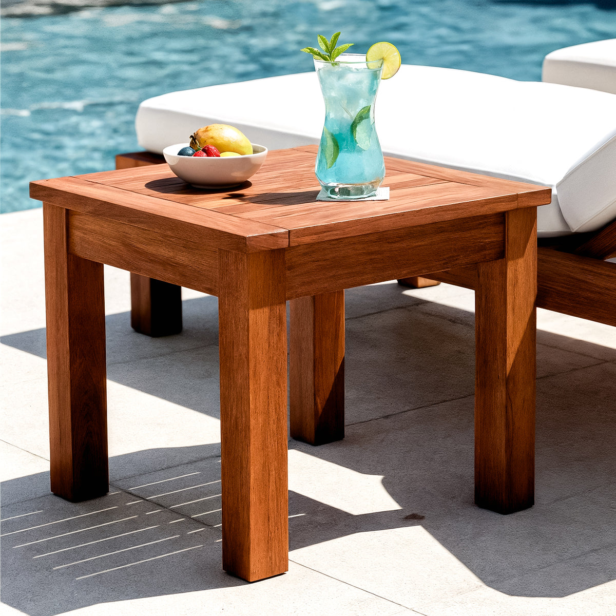 Laurette Outdoor Timber Side Table in Natural, placed poolside, holds a white bowl of fruit and a glass of blue drink, embodying summer relaxation with its durable, FSC Eucalyptus design.