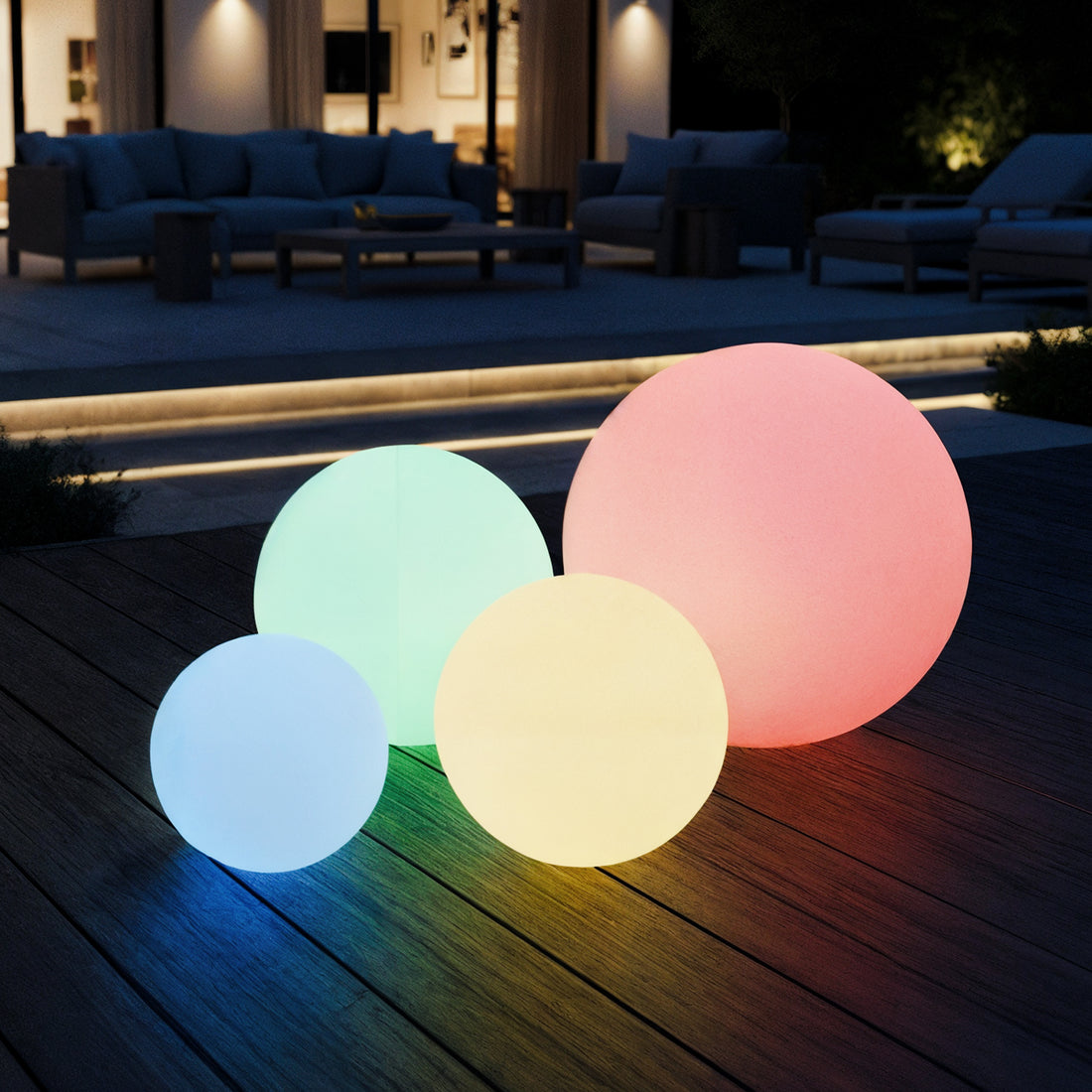 Four Sphero 50cm Floating Waterproof Outdoor LED Ball Lights in various sizes rest on an outdoor deck, creating an enchanting atmosphere with a modern seating area visible in the background.