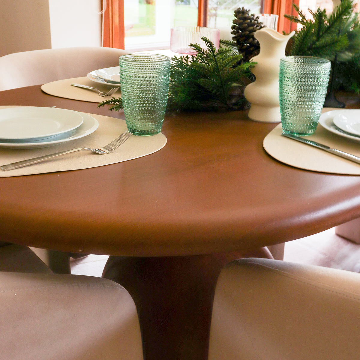 Close-up of the Lumen 220cm Oval Outdoor Concrete Dining Table in Chestnut, elegantly set with white plates, pale-green glasses, and an evergreen centerpiece, surrounded by plush cream-colored chairs.