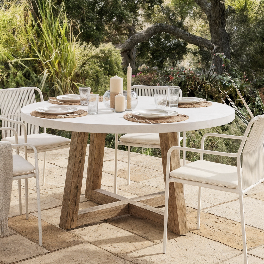 Magnus Hardwood and Concrete Outdoor Dining Table in White with a round tabletop and acacia wood base, surrounded by four metal-frame chairs with woven backs, set for dining.