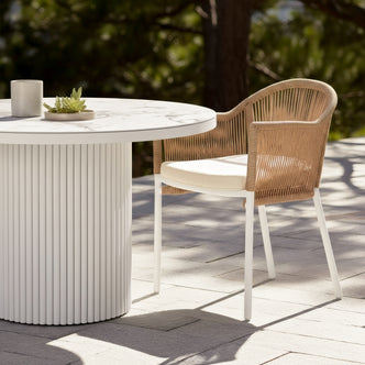 Mindanao PE Wicker Outdoor Dining Chair in Natural with beige cushion and white legs, shown beside a white fluted outdoor