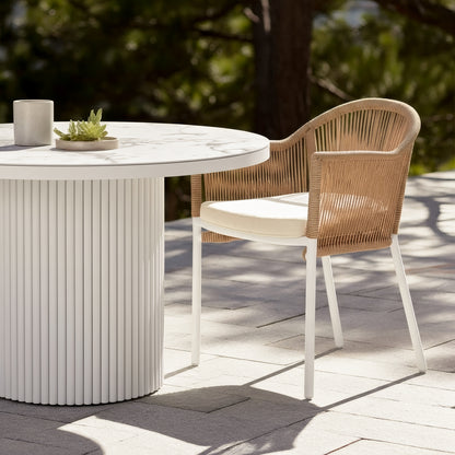 Mindanao PE Wicker Outdoor Dining Chair in Natural with beige cushion and white legs, shown beside a white fluted outdoor