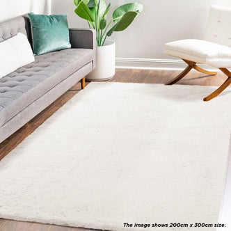 Azaliya 200x300cm Faux Rabbit Fur Floor Rug in Cream White, set in a modern living room with a gray button-tufted sofa, green velvet pillow, and a white cushioned chair, enhancing the room's elegance.