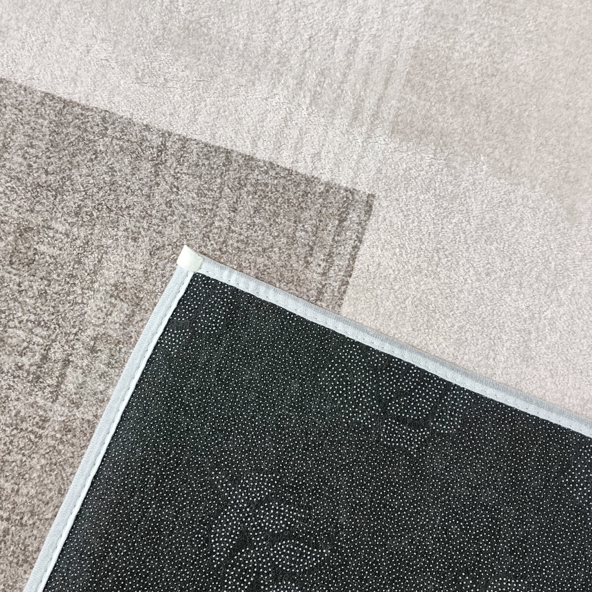 Close-up of the Orsa 200x300cm Small Contemporary Indoor Floor Rug, showing its corner flipped to reveal the black, dot-patterned non-slip backing and light fabric edge, resting on a beige carpet.