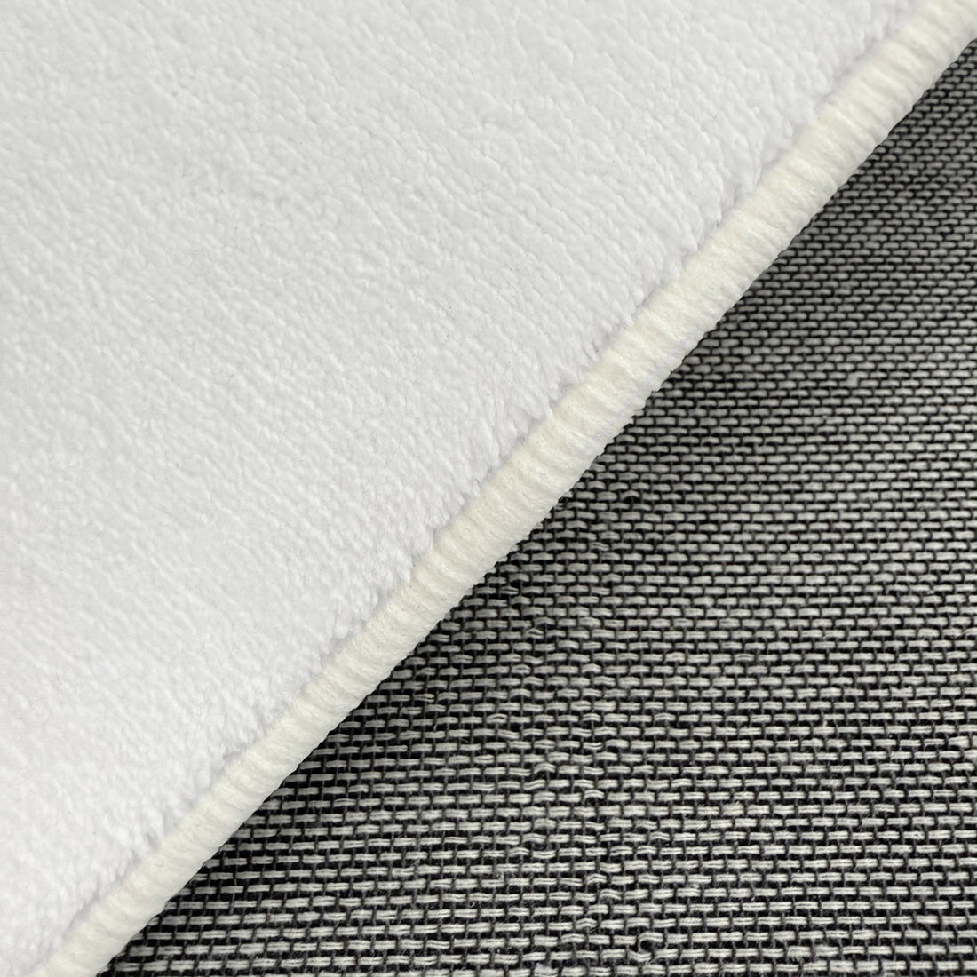 Close-up of Pixie 240x330cm Large Wavy Shag Rug in Cream White, showcasing its soft, fleece-like texture with a bound edge atop a woven grey-tone fabric background.