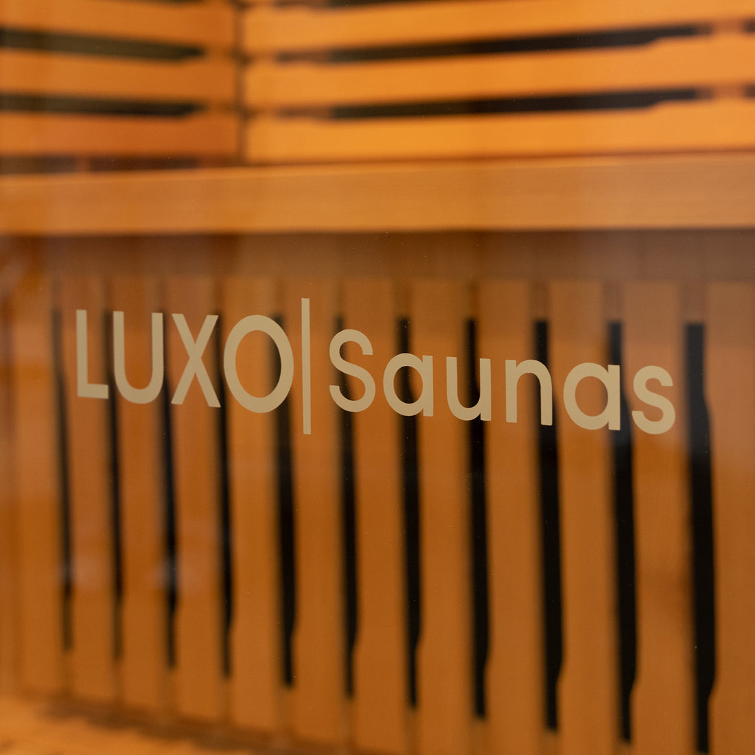 Harlow 1 Person Canadian Hemlock Infrared Home Sauna viewed through a glass door with LUXO | Saunas branding, showcasing wooden slatted benches and walls inside the sauna.