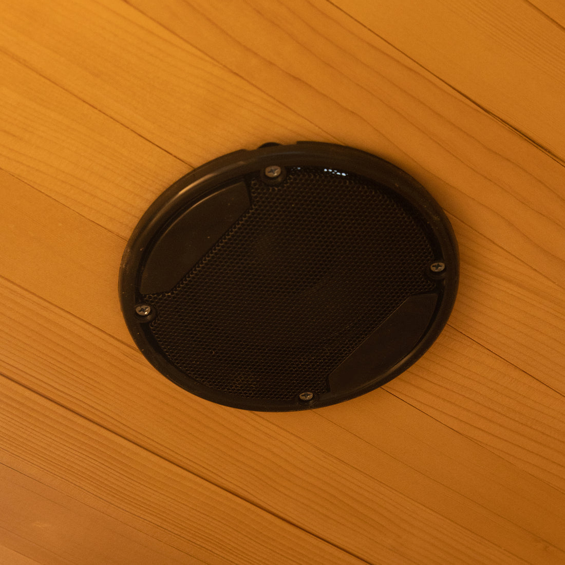 Circular black grille on wood ceiling, likely part of the Harlow 2 Person Canadian Hemlock Infrared Home Sauna, features built-in sound system and perforated metal cover, enhancing sauna experience.