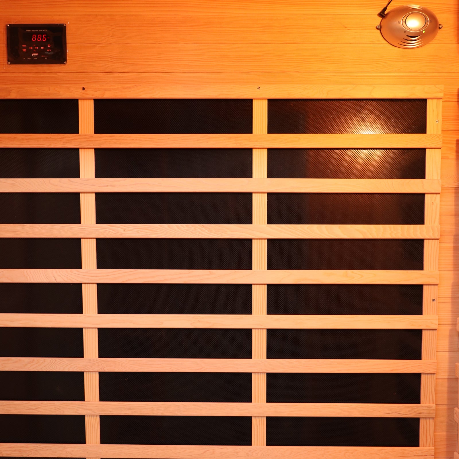 Valo 3 Person Carbon Fibre Infrared Indoor Sauna interior features a black heating panel behind wooden slats, digital control near the ceiling, and a light or speaker fixture, offering luxurious home relaxation.