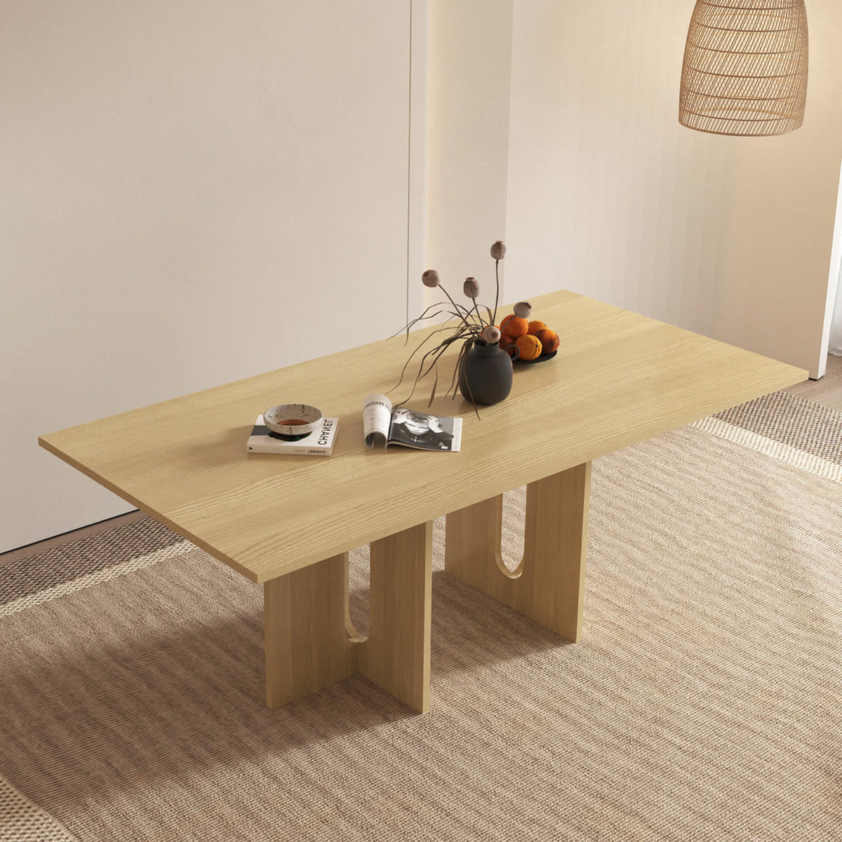 Amora 200cm Dining Table - Natural, featuring a light-wood top and sculptural legs with U-shaped cutouts, adorned with reading materials, a black vase, and a bowl of oranges.