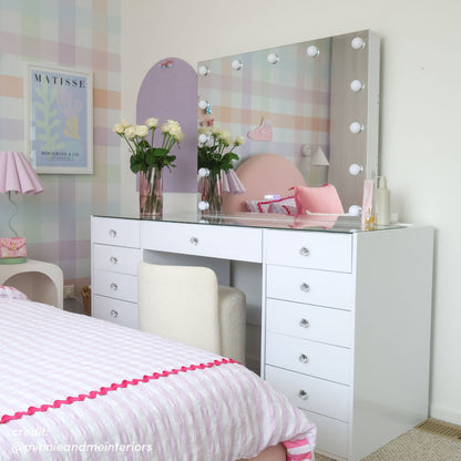 White makeup vanity with Hollywood LED mirror, glass top, crystal drawer knobs, and white boucle stool in a bedroom setting