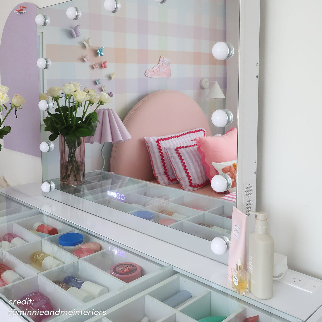 White Hollywood vanity with LED bulbs, open storage drawers with cosmetics, reflecting pink bedhead and checkered wall