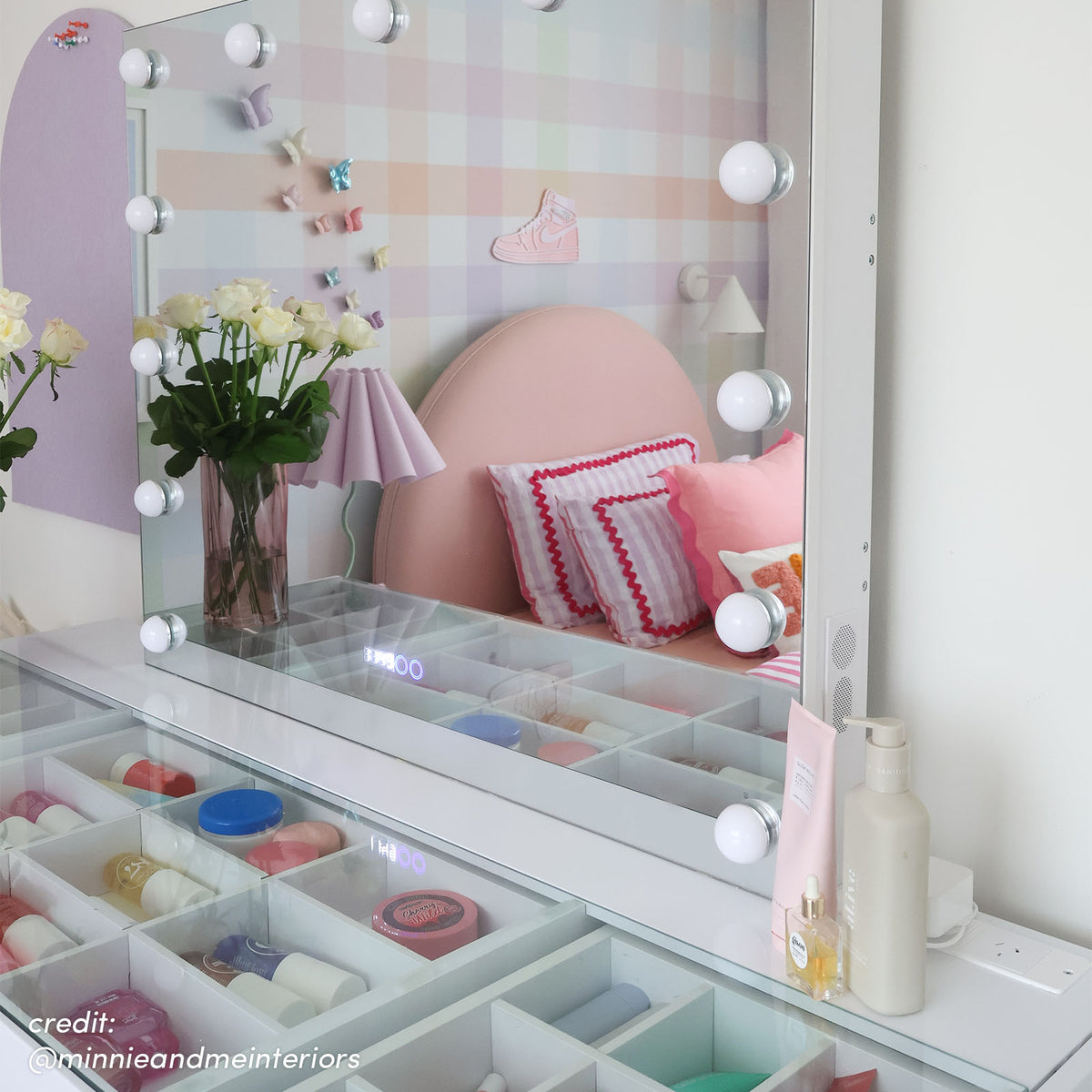White Hollywood vanity with LED bulbs, open storage drawers with cosmetics, reflecting pink bedhead and checkered wall
