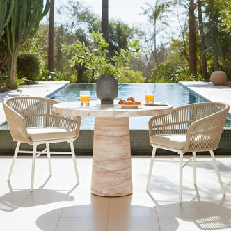 Round travertine concrete dining table with two rope chairs by pool, set with orange juice and pastries