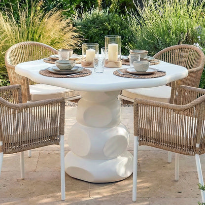 Azalea 110cm round white concrete outdoor dining table with sculptural base, set with plates, glasses and candles, surrounded